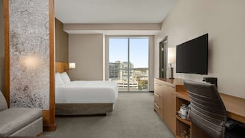 A hotel room with a large bed, a desk with a flat-screen TV, a chair, a dresser, and a large window offering a city view.