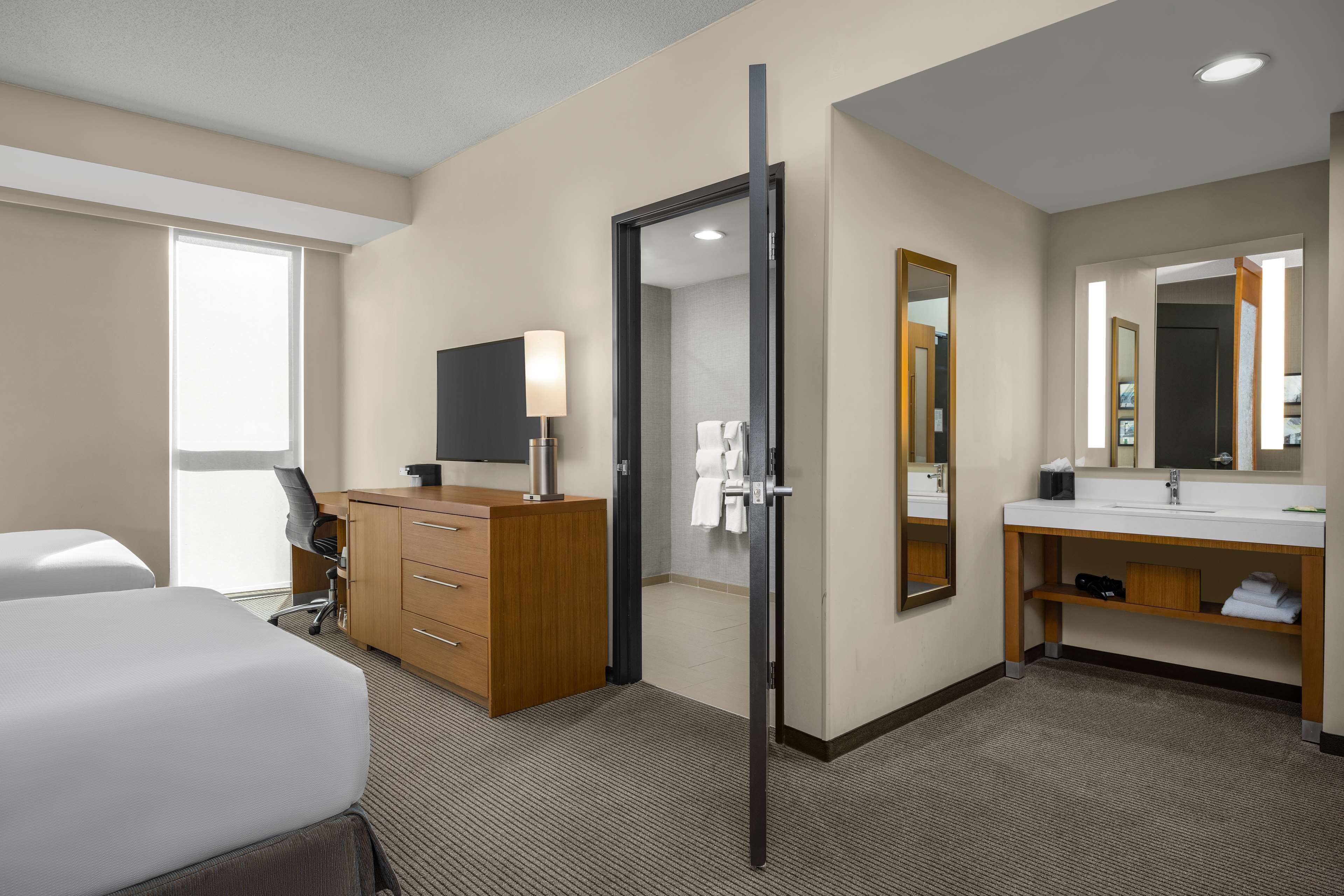 Une chambre d’hôtel comprenant un lit, un bureau, une chaise, une commode, un miroir, un lavabo et une fenêtre.
