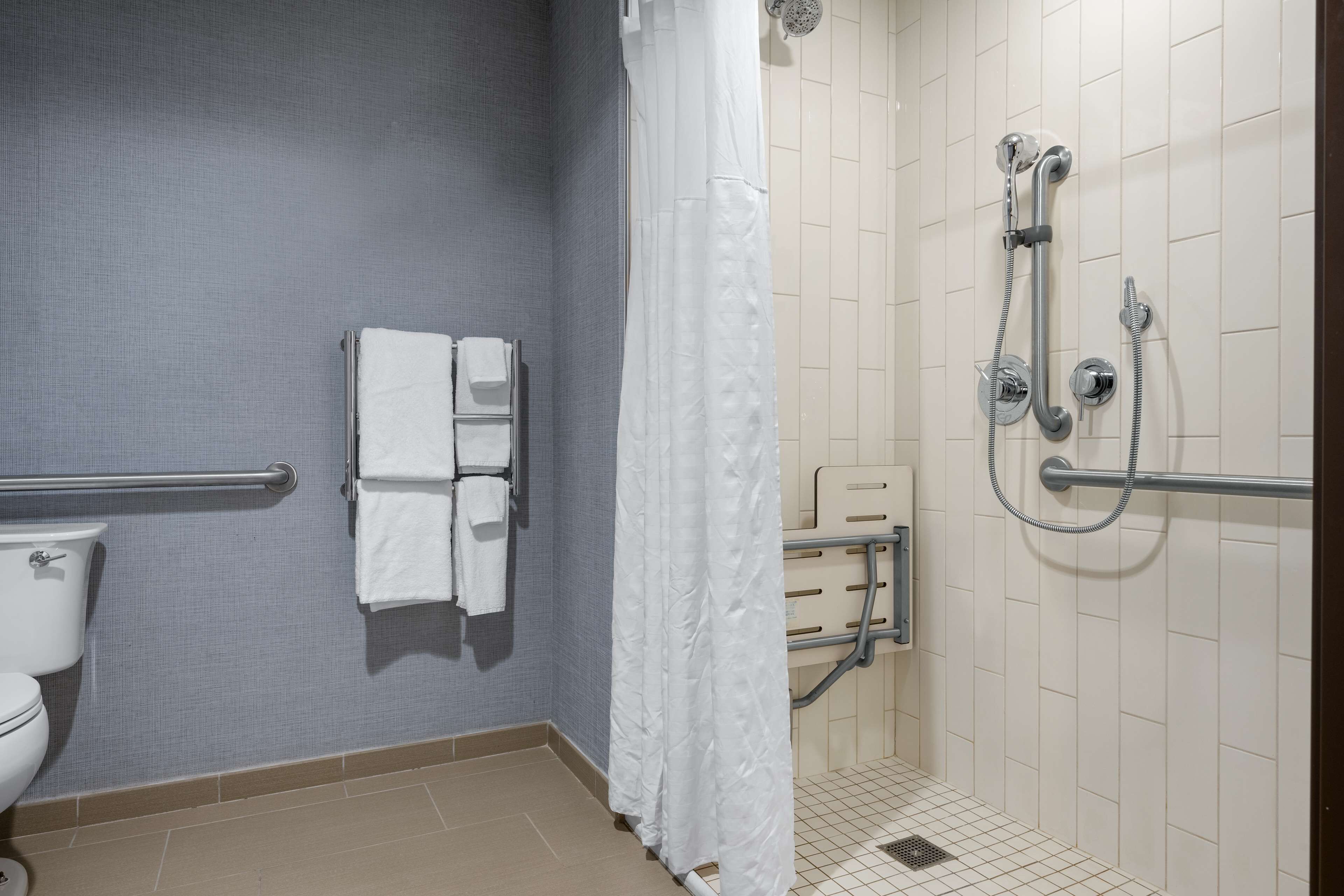 A bathroom with a toilet, a towel rack with towels, a shower with a handheld showerhead, and a grab bar.