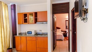A compact kitchen with wooden cabinets, a microwave, and a sink. A small trash can is visible near the entrance. - Nobilis Apartments (Kigali)
