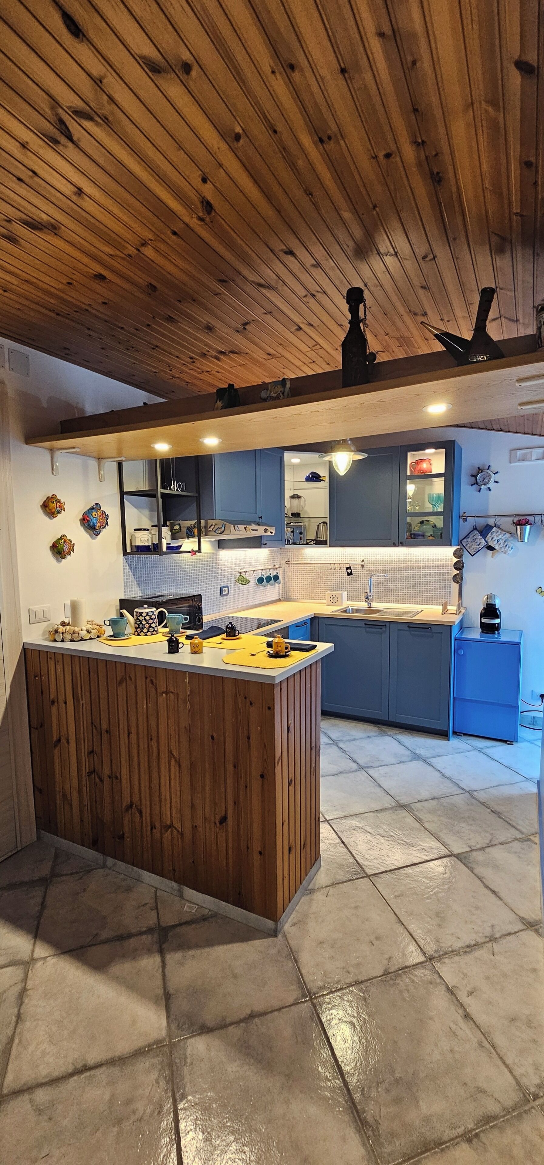 A kitchen with wooden ceiling beams, a central island, blue cabinets, and a tiled floor.