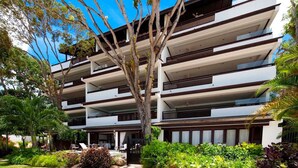 A multi-story hotel with balconies, surrounded by lush greenery and a clear blue sky. - Coral Cove 11 by Altman (Paynes Bay)