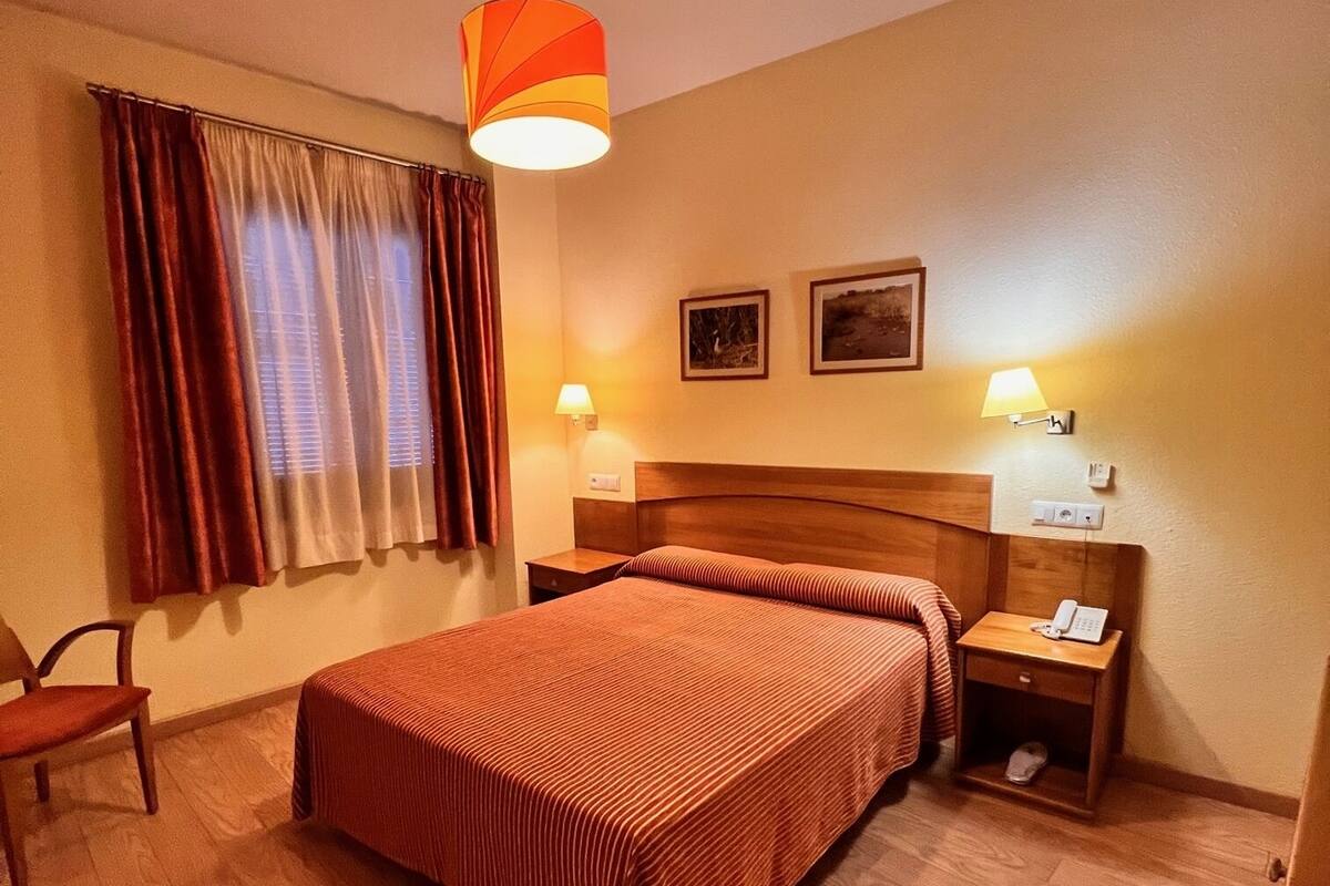 A hotel room with a wooden floor, a bed with striped bedding, a nightstand with a lamp, and a chair.