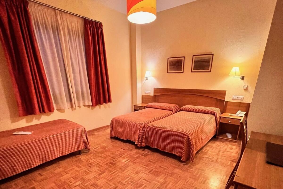 A hotel room with two beds, a desk, a chair, a lamp, and a window with curtains.