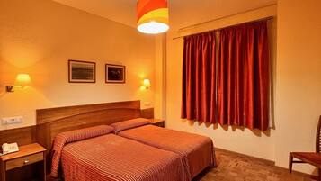 Una habitación de hotel con suelo de madera, una cama doble con ropa de cama a rayas, dos mesitas de noche con lámparas, un cabecero de madera, una cortina roja y una lámpara colgante.