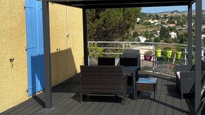 A covered outdoor seating area with wicker furniture, a pergola, and a view of a residential area. - La Maison Provençale (Unieux)