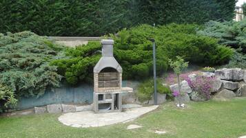 Un horno de piedra al aire libre en un jardín con pared de piedra, arbustos verdes y un árbol pequeño.