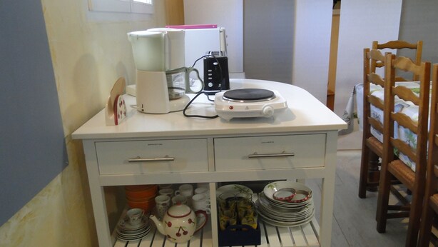 A kitchen with a white countertop, a coffee maker, a kettle, and a stove. There are shelves below holding dishes and a frying pan. - La Maison Provençale (Unieux)
