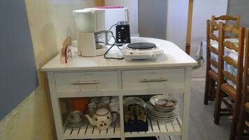 Een keuken met een wit aanrecht, een koffiezetapparaat, een waterkoker en een fornuis. Daaronder zijn planken met servies en een koekenpan.