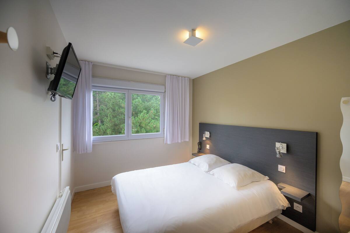 A bedroom with a bed, a television mounted on the wall, a window with curtains, and a nightstand with a lamp.