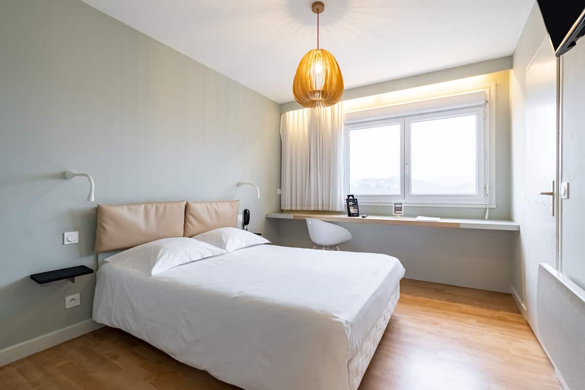 A bedroom with a bed, bedside table, lamp, window, and a desk with a chair.