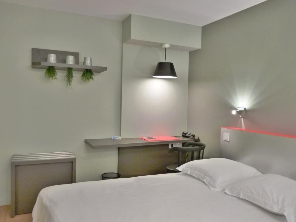 Una habitación de hotel moderna con una cama, un escritorio con una silla, un radiador pequeño, un estante con plantas decorativas y una lámpara de pared.