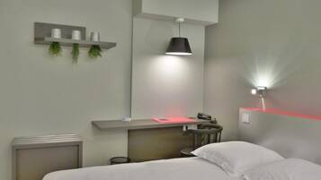 Una habitación de hotel moderna con una cama, un escritorio con una silla, un radiador pequeño, un estante con plantas decorativas y una lámpara de pared.