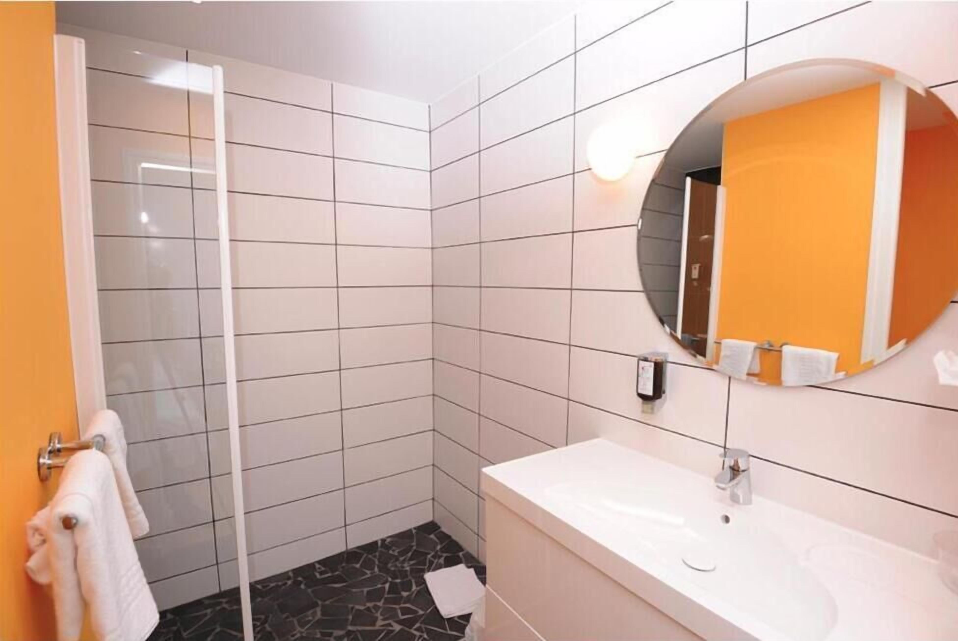 A bathroom with white tiled walls, a black and white patterned floor, a white sink, and a round mirror with an orange towel hanging on it.