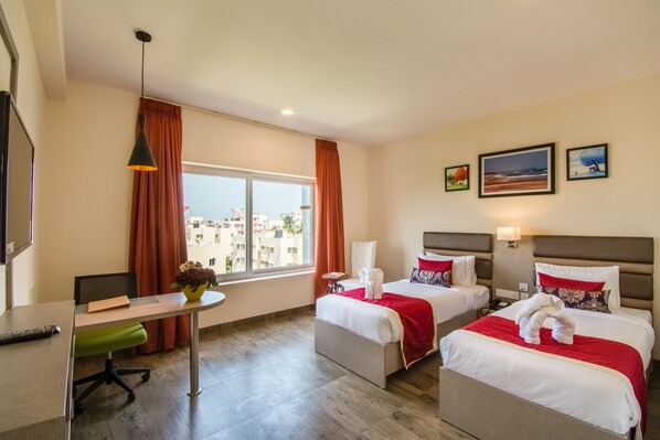 A hotel room with two beds, a desk, a chair, a small table, and a window with curtains. - Mantris Hotel (Visakhapatnam)