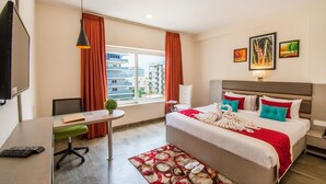 Quarto de hotel com cama, escrivaninha, cadeira, mesinha, abajur e janela com cortinas.