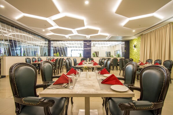 A spacious dining area with hexagonal ceiling lights, black upholstered chairs, and neatly arranged tables with red napkins and silverware. - Mantris Hotel (Visakhapatnam)