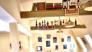 A spacious room with a high ceiling, featuring a dining area with multiple tables and chairs, a bookshelf with various books, and a wall adorned with framed pictures. - Old Town Mtatsminda (Tbilisi)