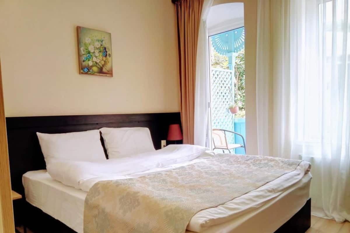 A neatly made bed with white linens and a patterned blanket, a wooden headboard, a painting on the wall, and a view of the outside through a window with curtains.