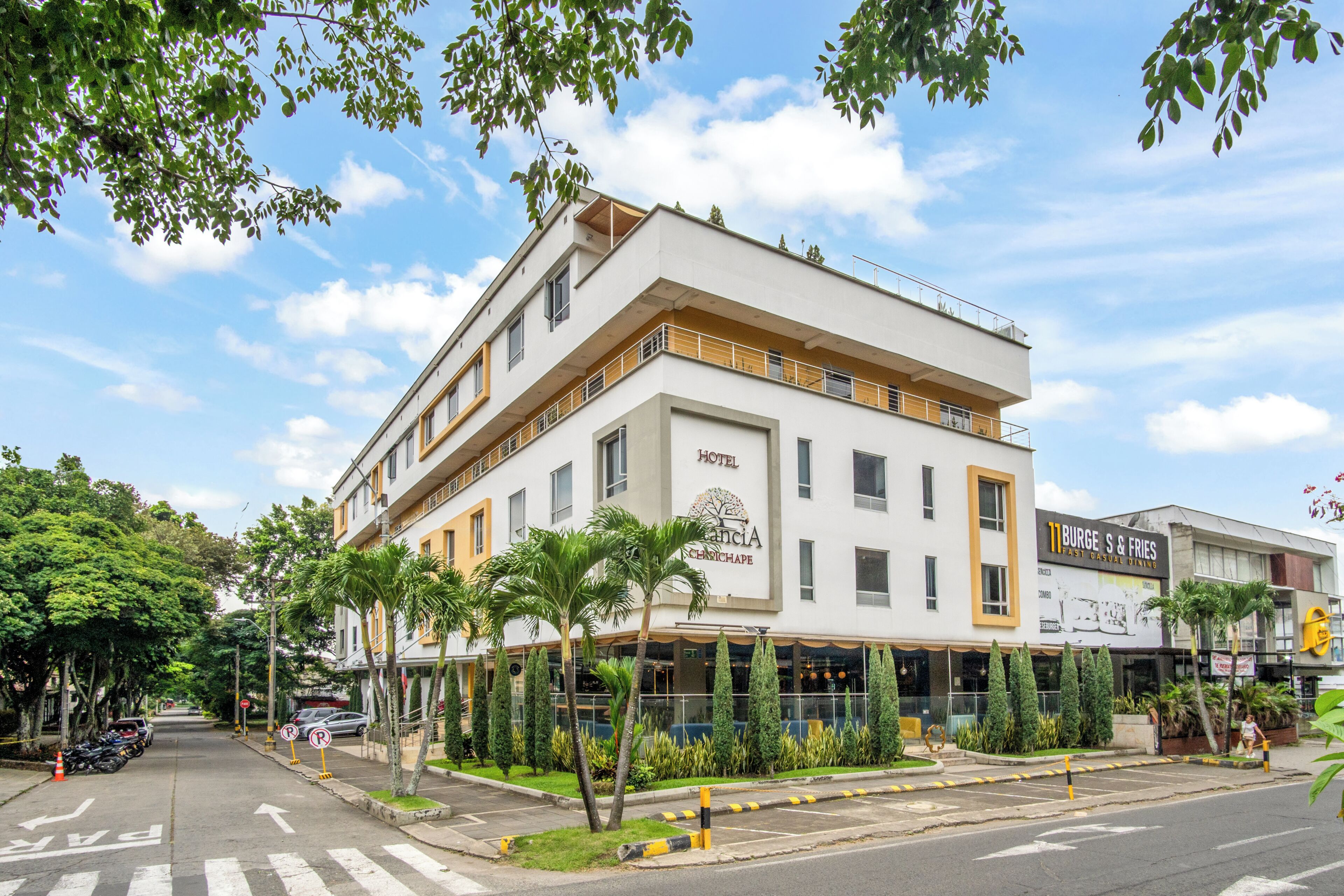 A hotel with a modern design, a visible restaurant named '11 BURGE S & FRIES,' and a street view with parked cars and greenery.