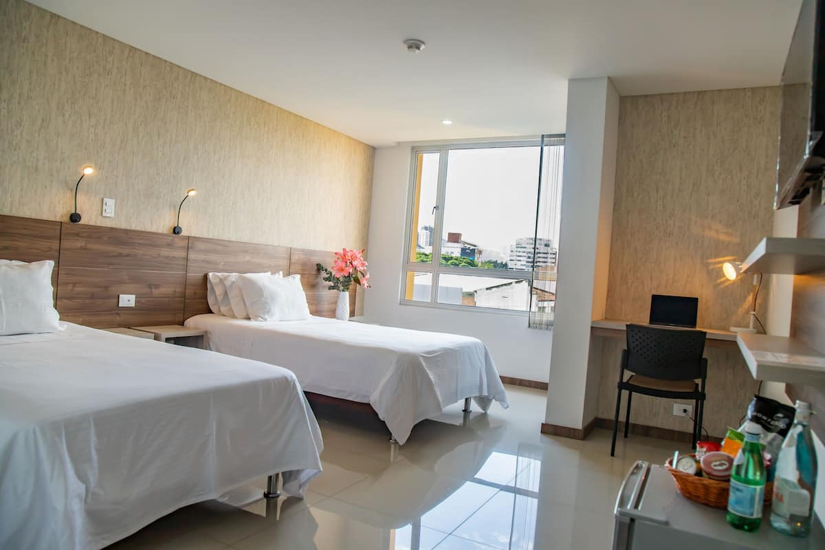 A hotel room with two beds, a desk, a chair, a window with a view, and a small kitchenette area.