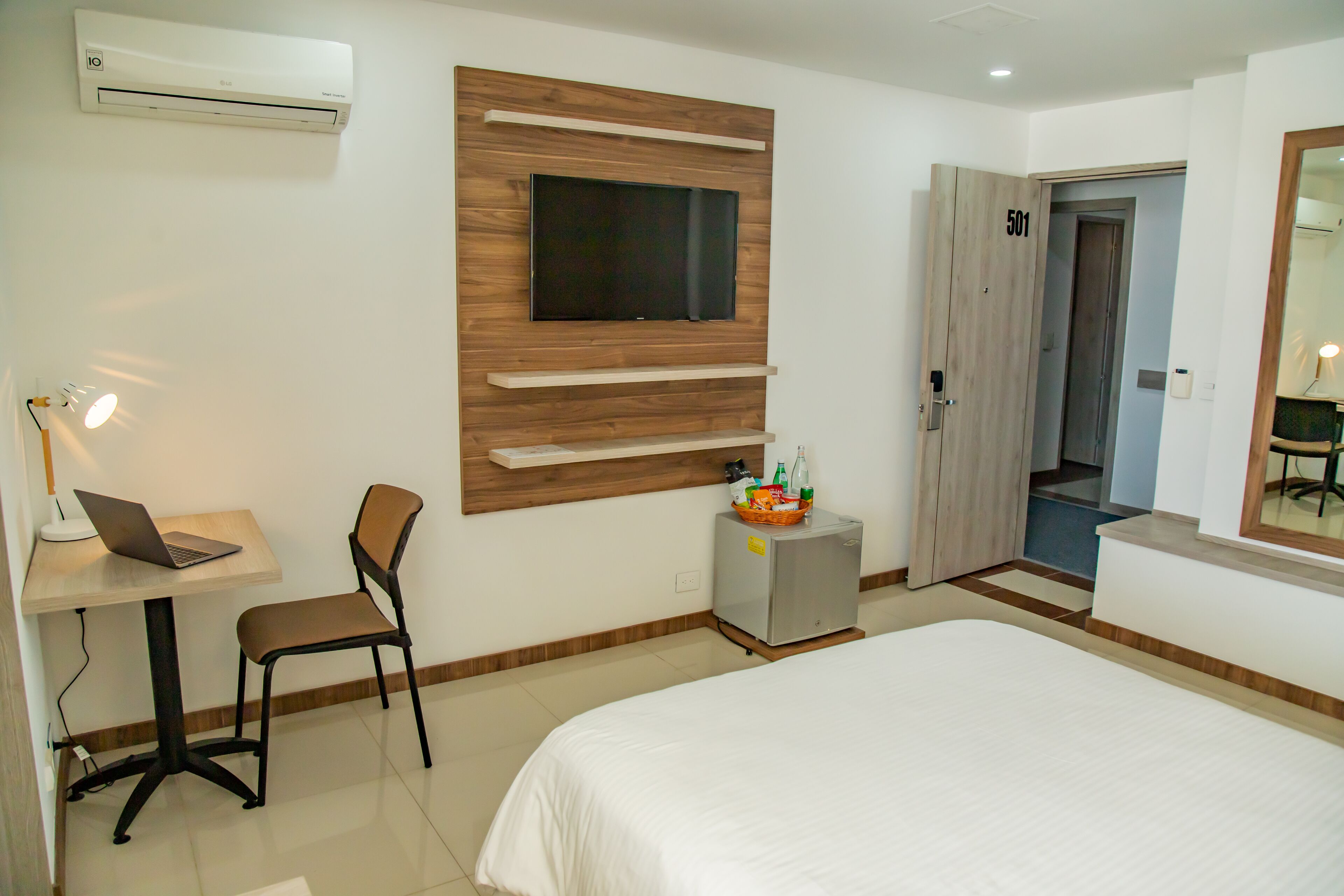 A hotel room with a bed, a desk with a laptop, a chair, a TV mounted on the wall, a minibar, and a door to another room.