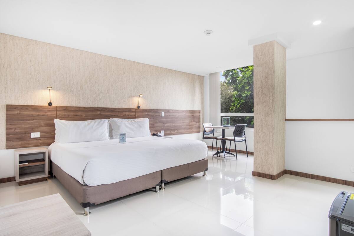 A modern hotel room with a large bed, bedside tables, a desk with chairs, and a view of greenery outside.