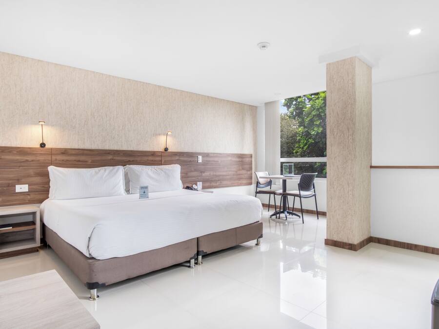 Habitación de hotel moderna con una cama grande, mesitas de noche, un escritorio con sillas y vistas a zonas verdes.