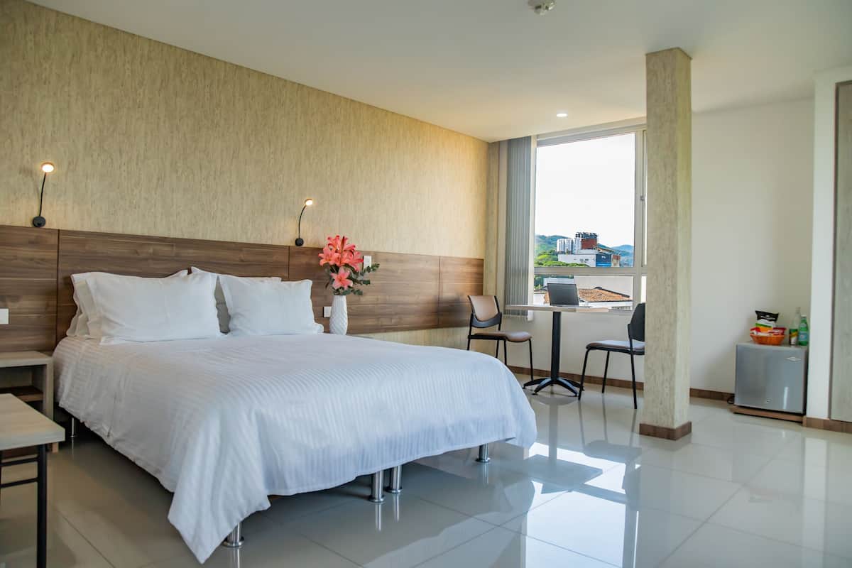 A hotel room with a large bed, a desk, a chair, a small table, and a window with a view of buildings.