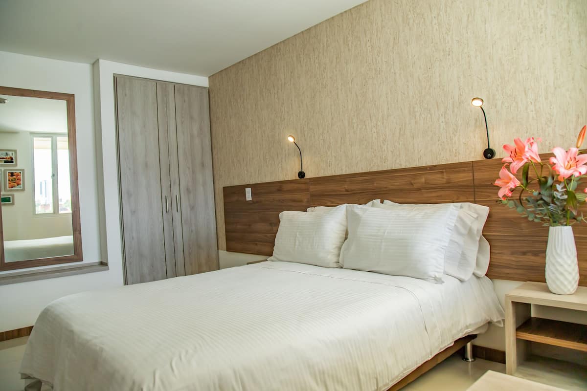 A modern bedroom with a wooden headboard, a large bed with white linens, a nightstand with a vase of flowers, and two wall-mounted lights.