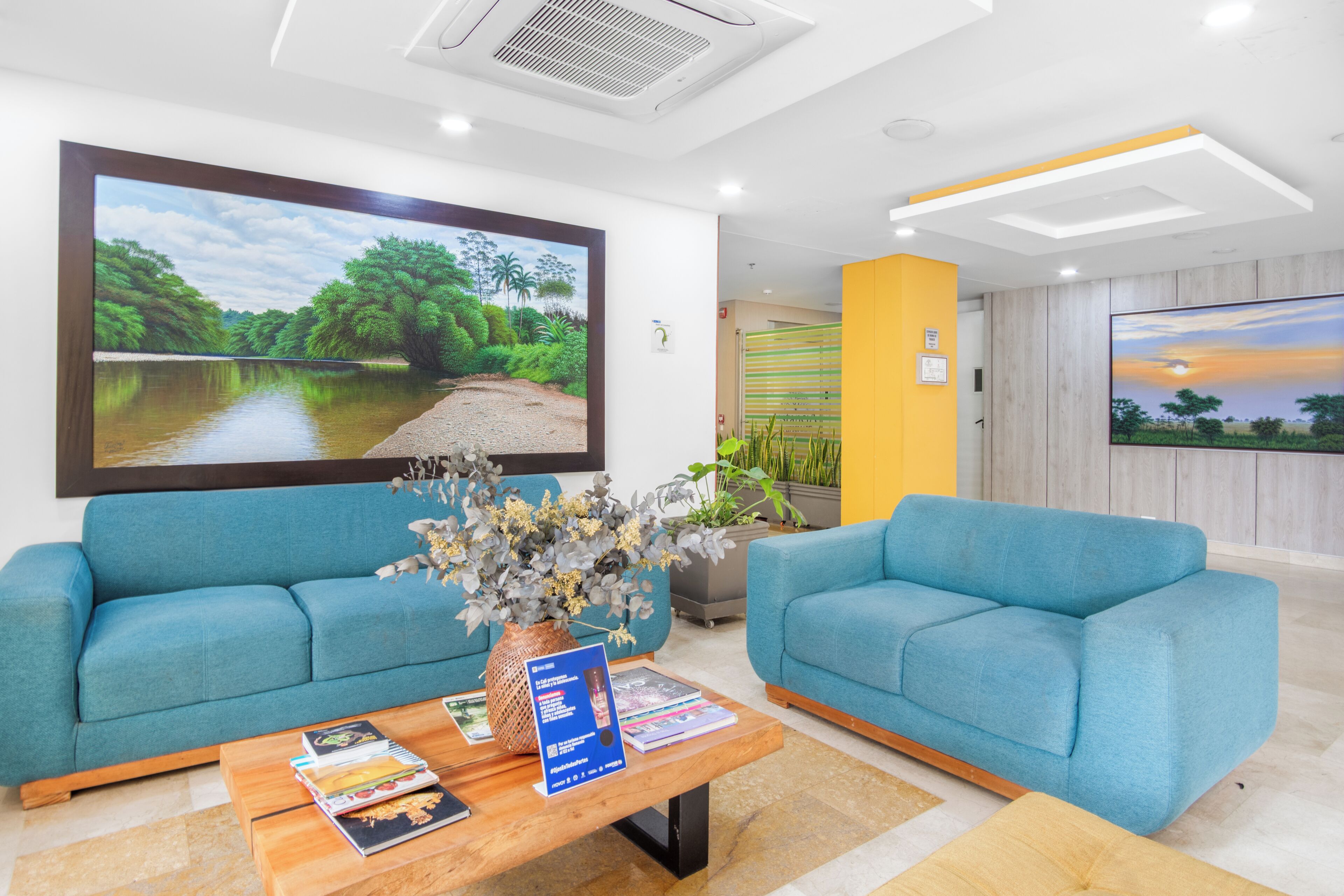 A reception area with two blue sofas, a wooden coffee table with magazines, a large framed landscape painting, and a TV screen displaying a sunset.