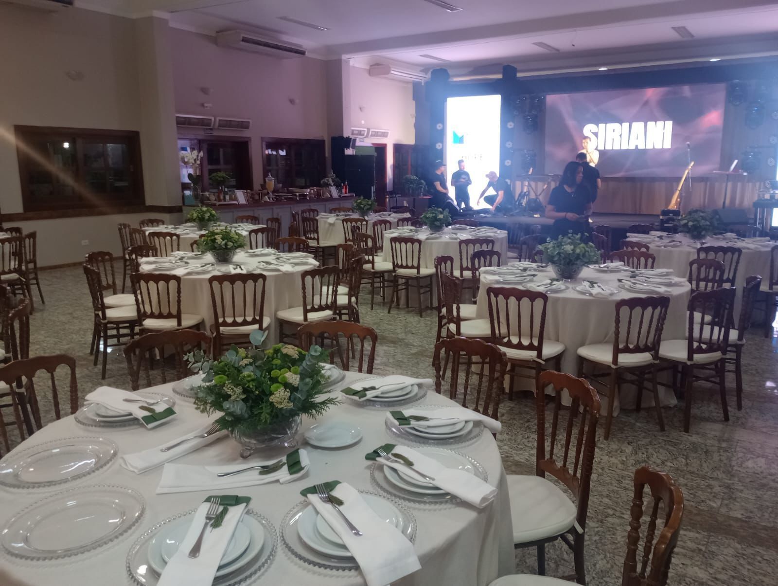 Una sala da banchetto con tavoli rotondi apparecchiati per un evento formale, con tovaglie bianche, tovaglioli verdi e bicchieri trasparenti.
