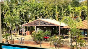 Zona de piscina con área de descanso cubierta, plantas en macetas y vistas al entorno verde.