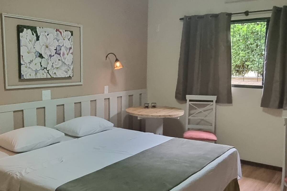 Ein Hotelzimmer mit einem Bett, zwei Nachttischlampen, einem kleinen runden Tisch, einem Sessel, einem Bild an der Wand und einem Fenster mit Vorhängen.