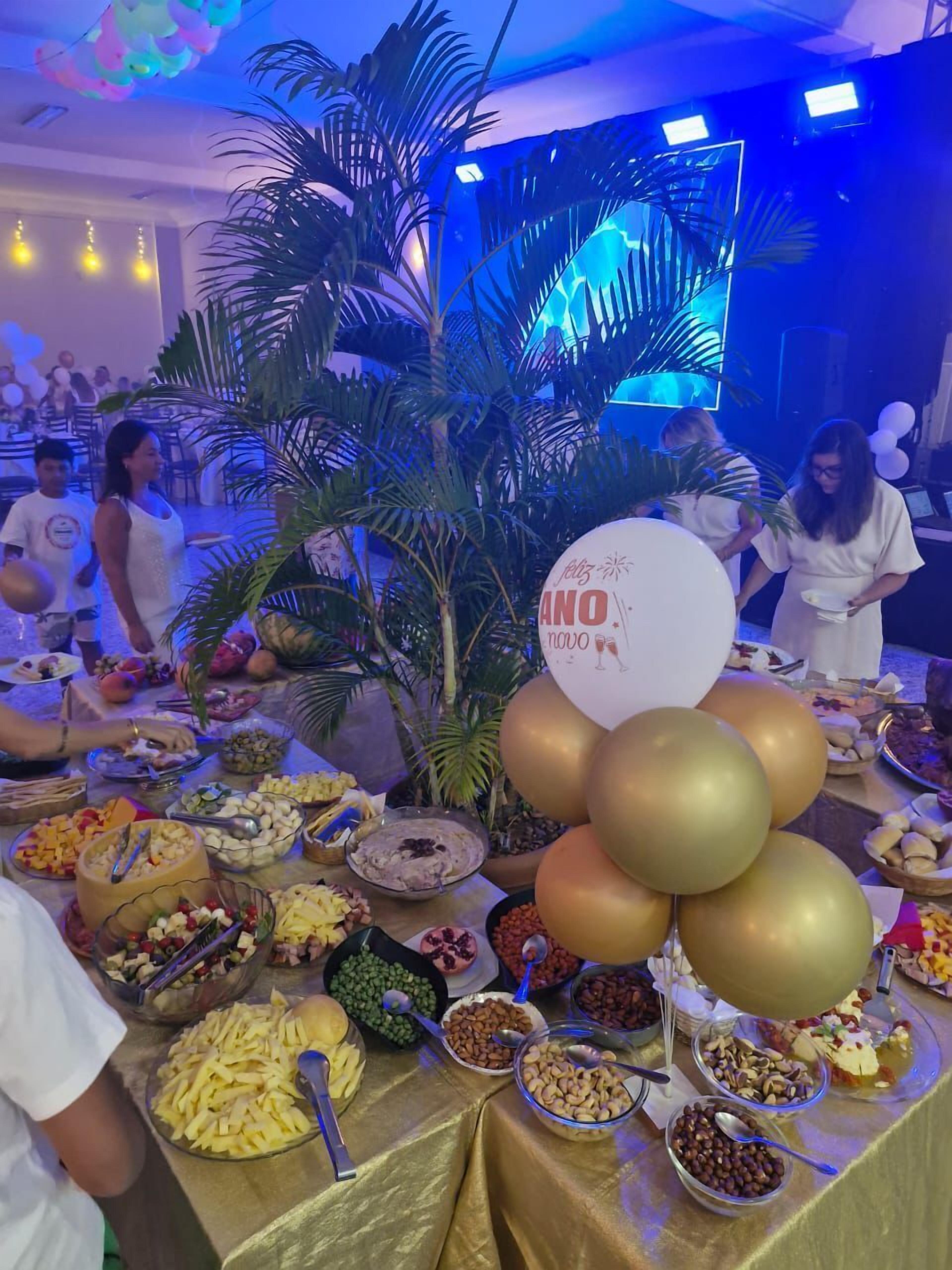 A buffet table with various food items and balloons, surrounded by palm plants and a blue-lit background.