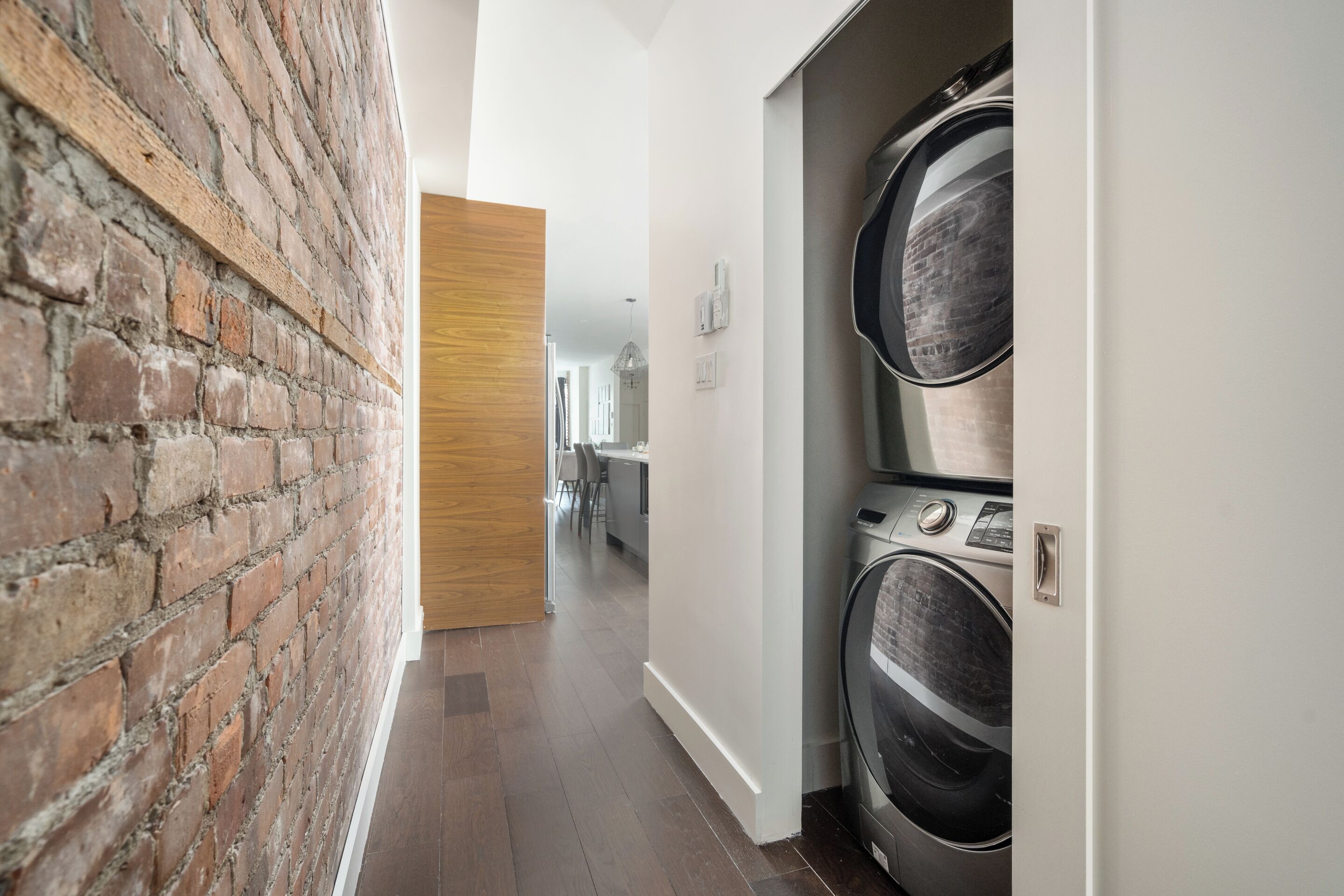 A laundry room with stacked washers and dryers, a brick wall, and a hallway leading to a kitchen area.
