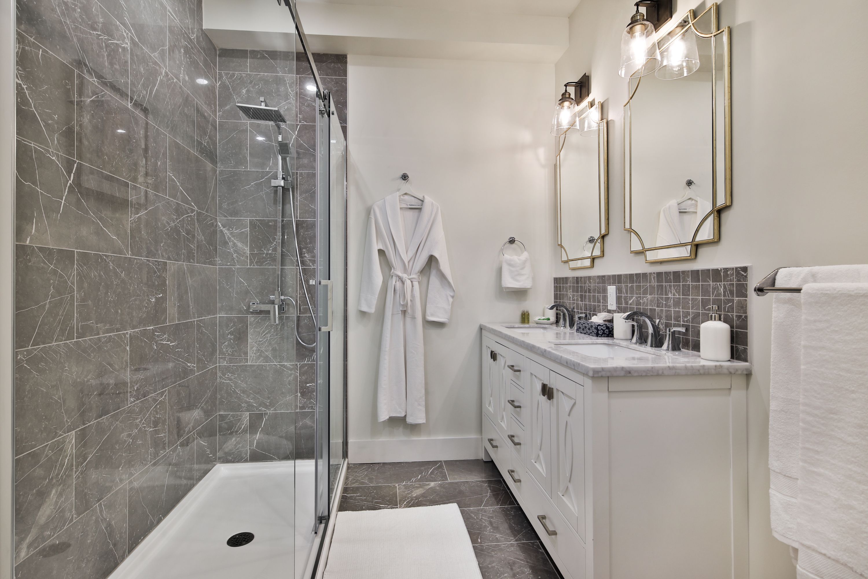 Un bagno moderno con cabina doccia in vetro, un mobile lavabo bianco con piano in marmo e due specchi a parete con cornice dorata.