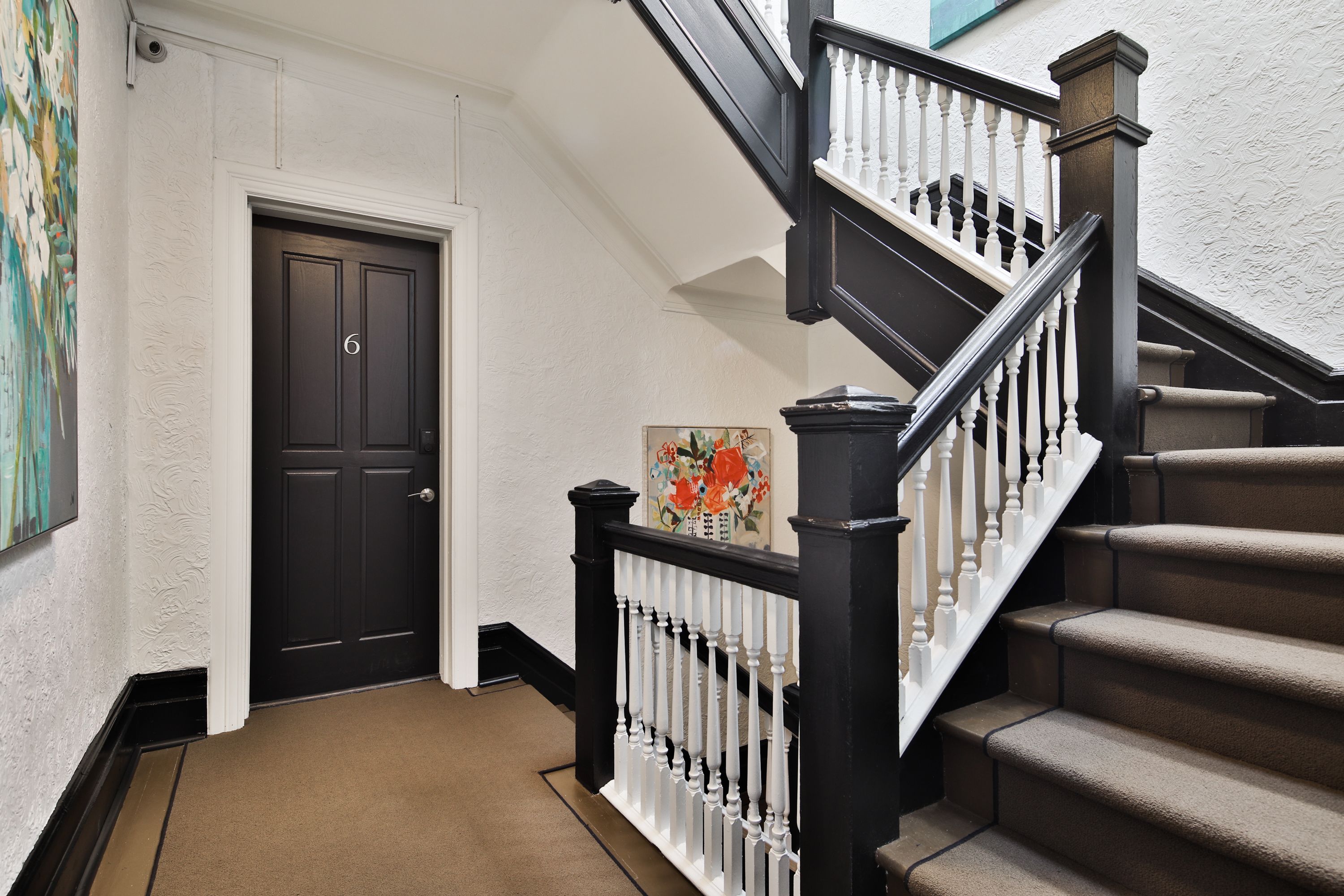 A hallway with a black door marked with the number 6, a staircase with white balusters, and a painting on the wall.
