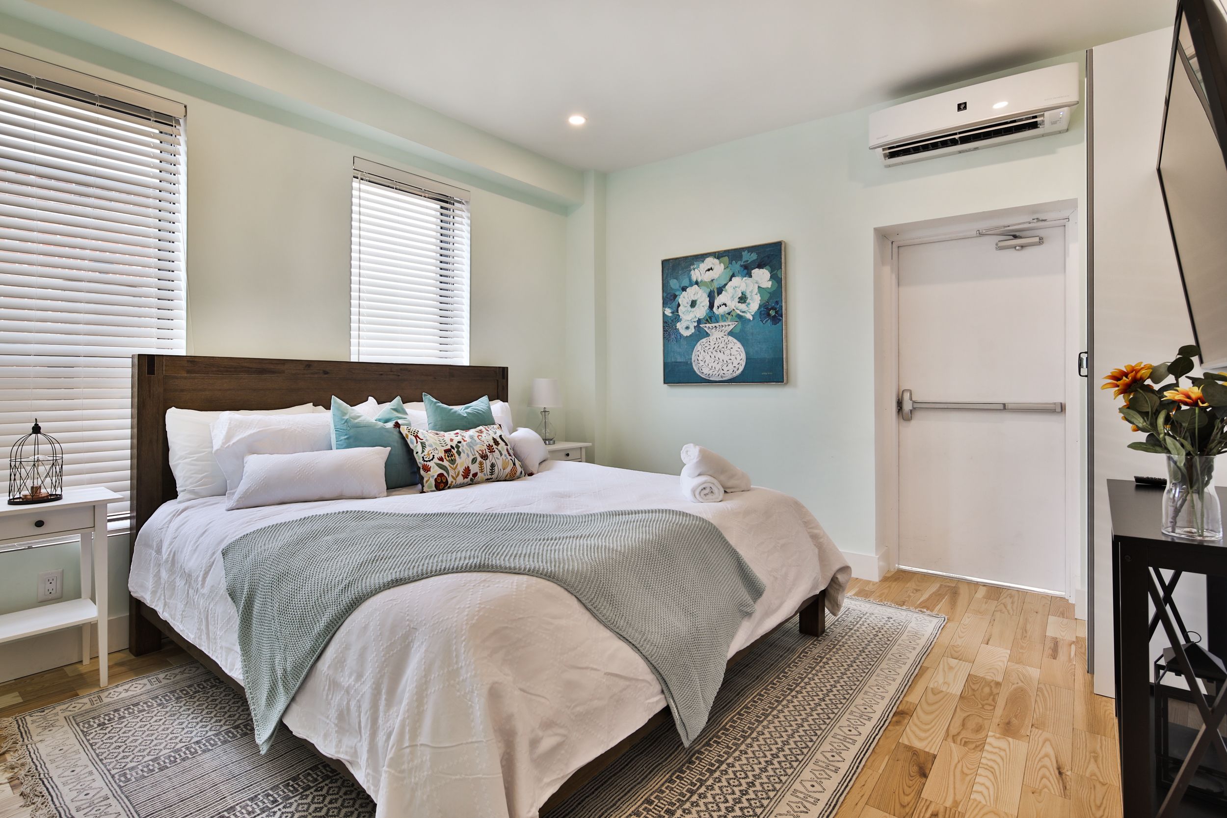 A bedroom with a bed, bedside tables, a nightstand, a wall-mounted air conditioner, and a door.
