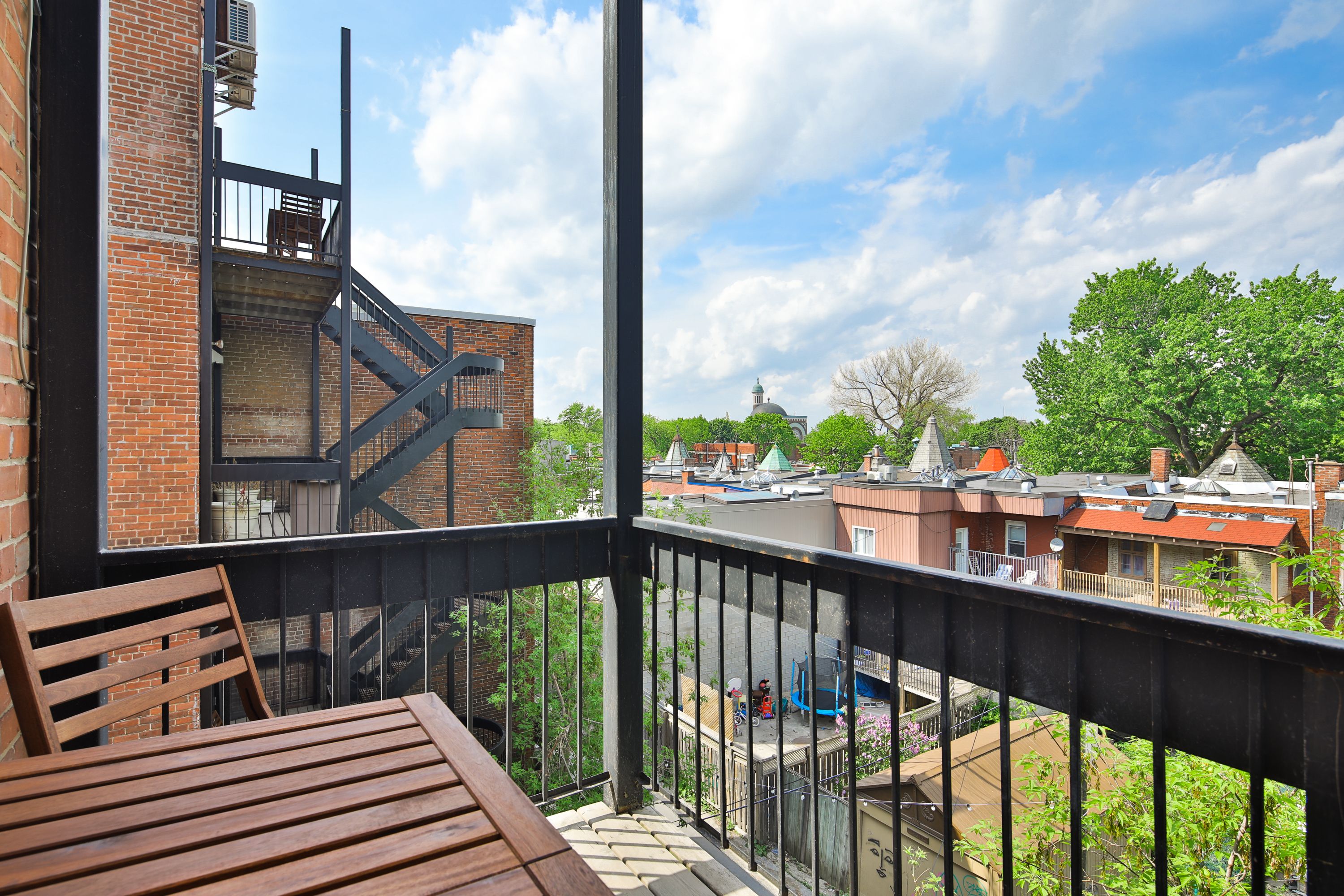 A balcony with a wooden table and chairs, overlooking a residential area with brick buildings and a church steeple.