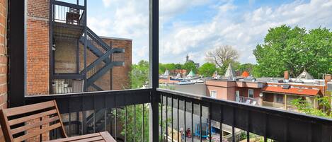 A balcony with a wooden table and chairs, overlooking a residential area with brick buildings and a church steeple.