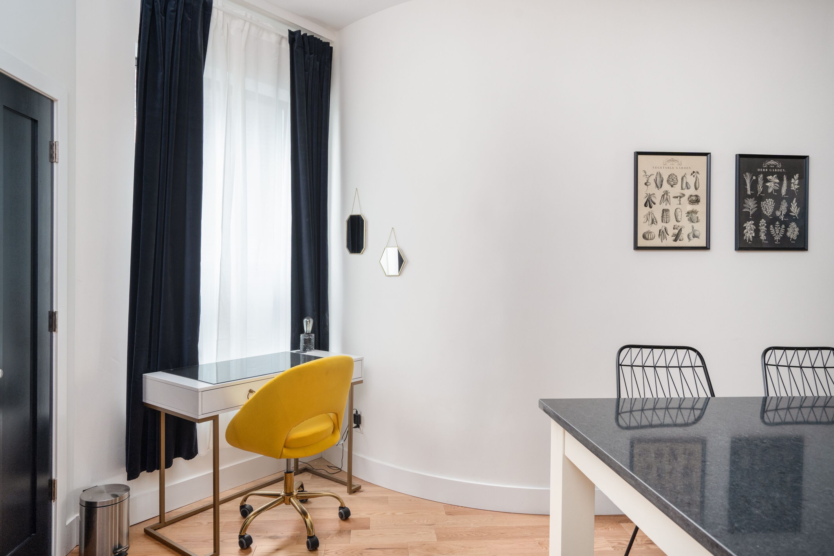A modern room with a glass desk, a yellow chair, a black dining table, and two framed botanical prints on the wall.
