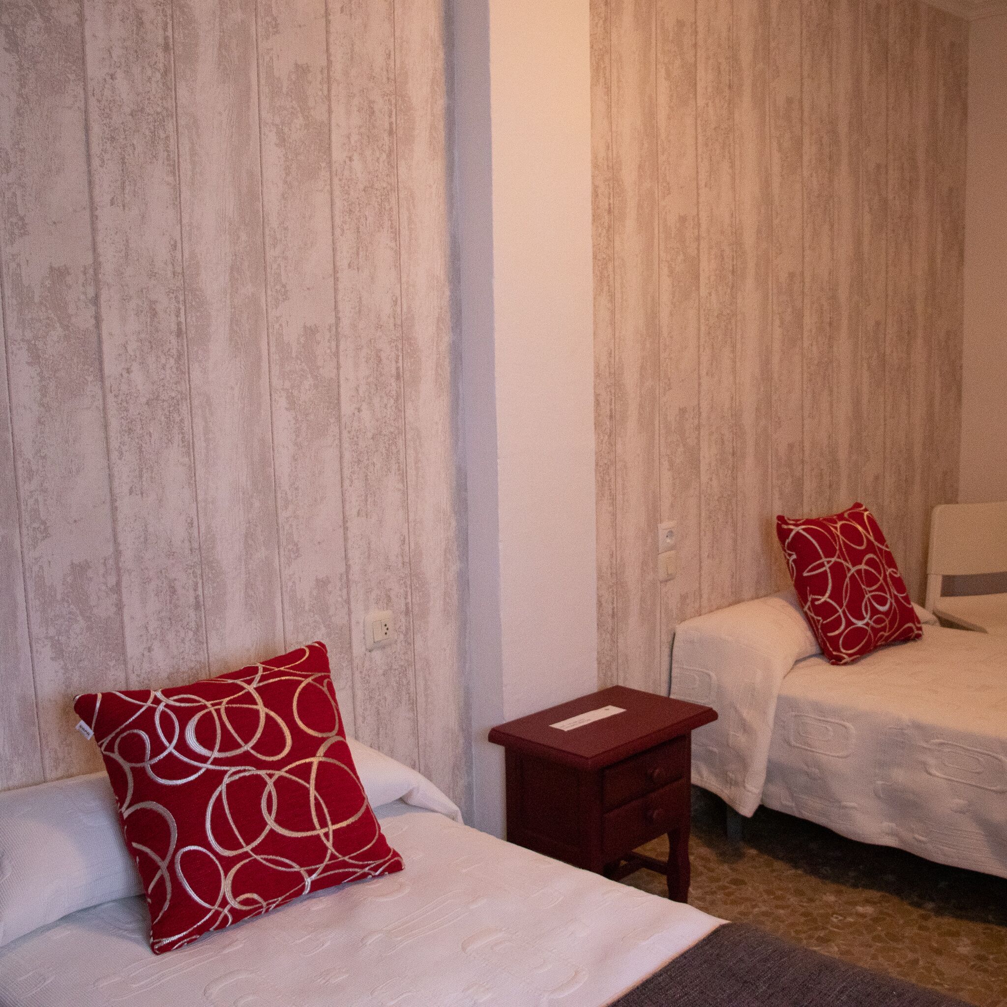 A hotel room with two beds, each with a red pillow, a small wooden nightstand, and a white wall.