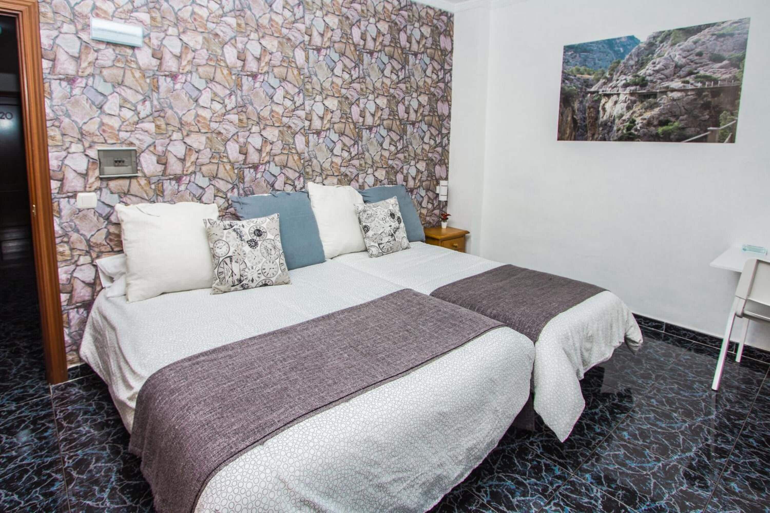 Een tweepersoonsbed met een muur erachter met een stenen patroon, een klein nachtkastje en een ingelijst beeld aan de muur.