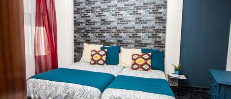 A double bed with patterned bedding, a brick-patterned headboard, a small bedside table with a plant, and a blue dresser.