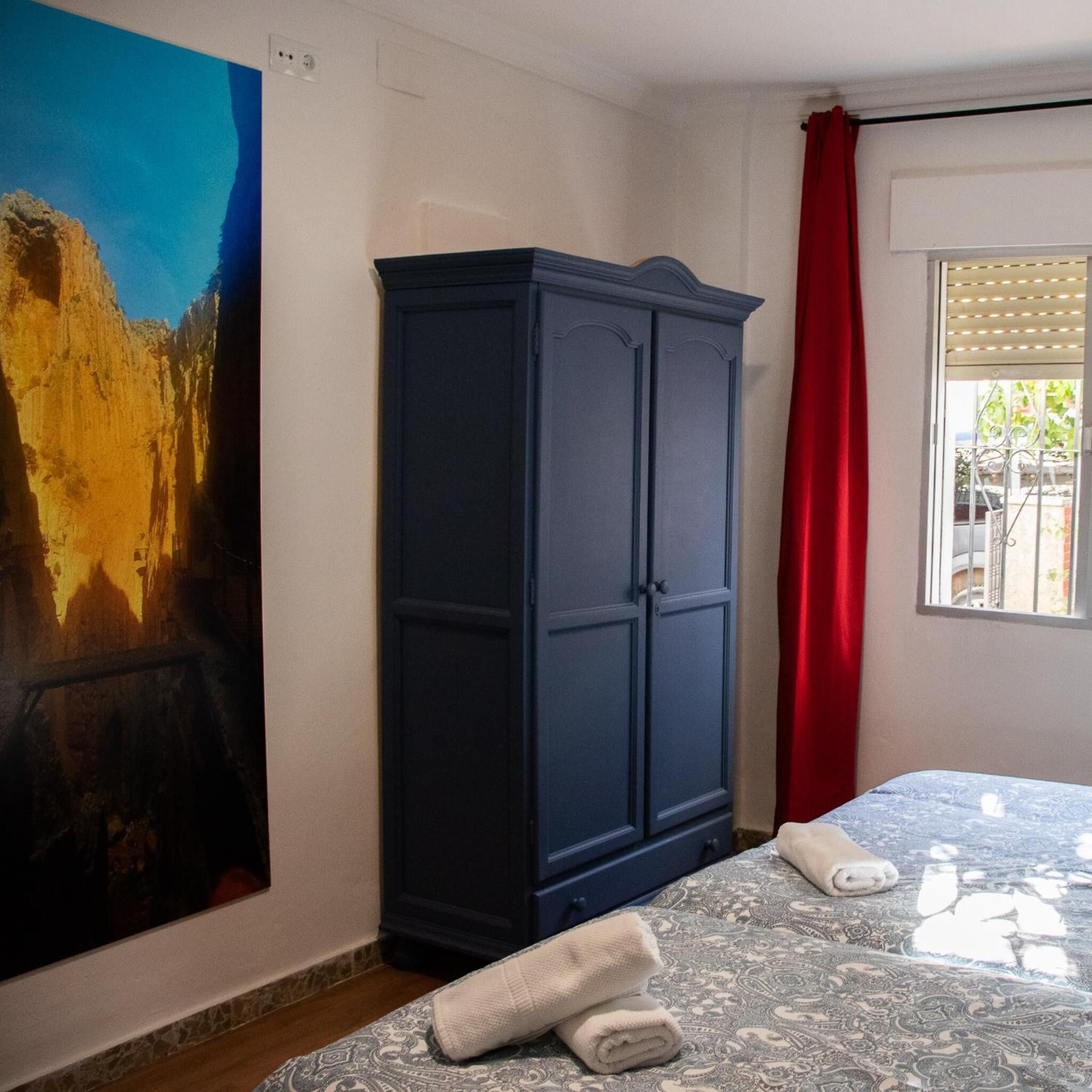 Um quarto com cama, um guarda-roupa azul, um quadro na parede e uma janela com cortina vermelha.