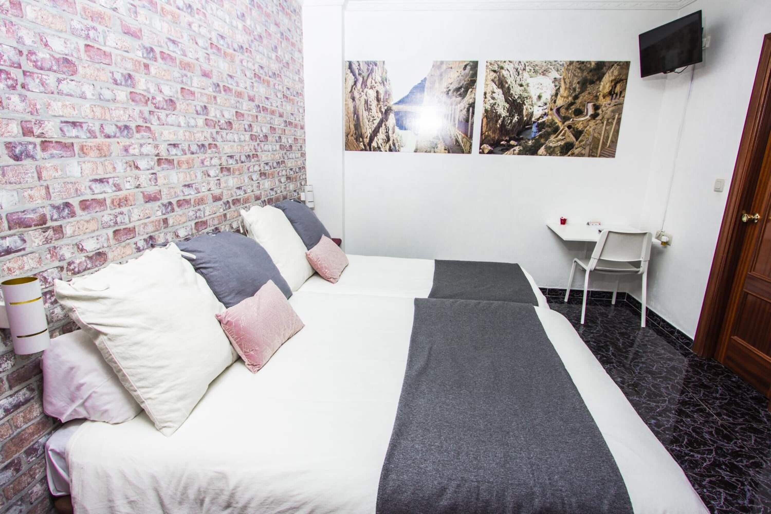 A hotel room with a brick-patterned wall, a bed with white and gray bedding, a desk with a chair, and a mounted TV.