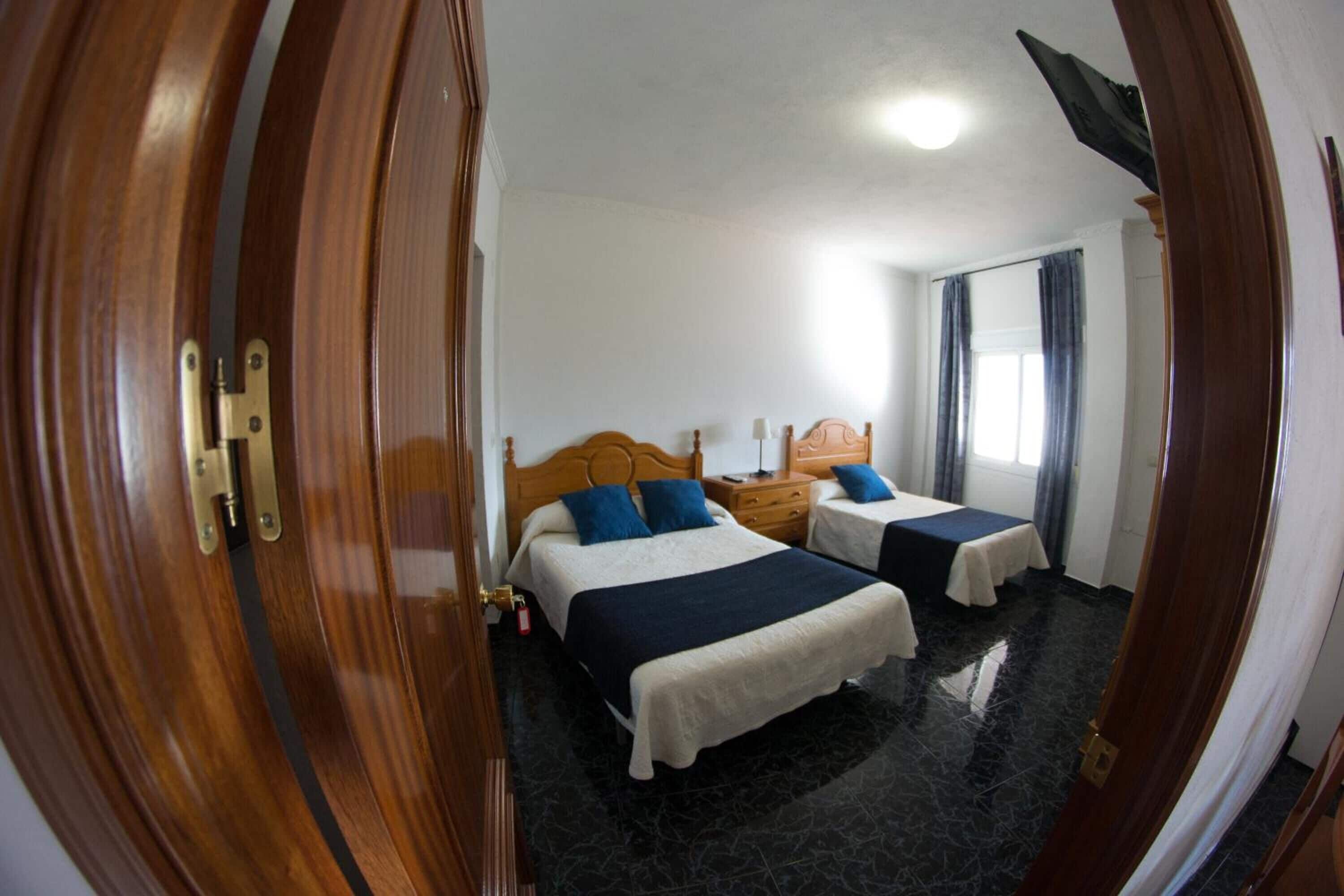 Habitación de hotel con dos camas, una puerta de madera, una mesita de noche, una lámpara y una ventana con cortinas.