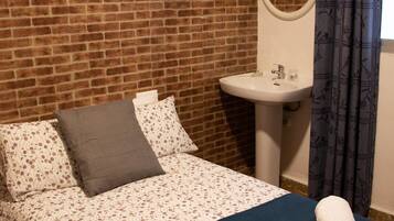 Una habitación con pared de ladrillo, una cama con ropa de cama floral, un lavamanos blanco y una cortina azul.