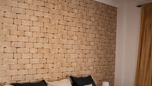 Una habitación de hotel con cabecera de ladrillo, cama con varias almohadas, mesita de noche con lámpara y ventana con cortinas.